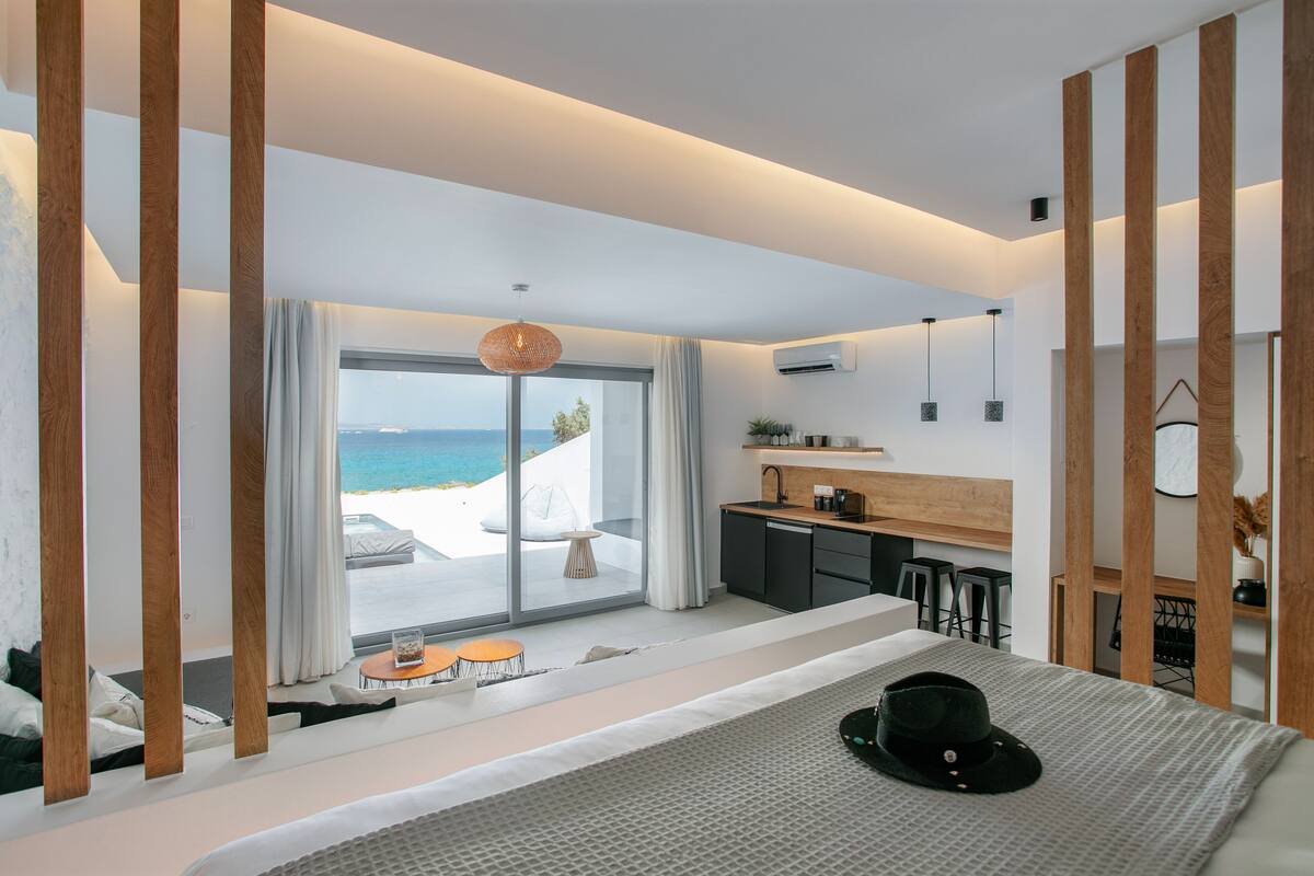 Habitación de hotel moderna con una cama grande, vista al mar, balcón con asientos y kitchenette.