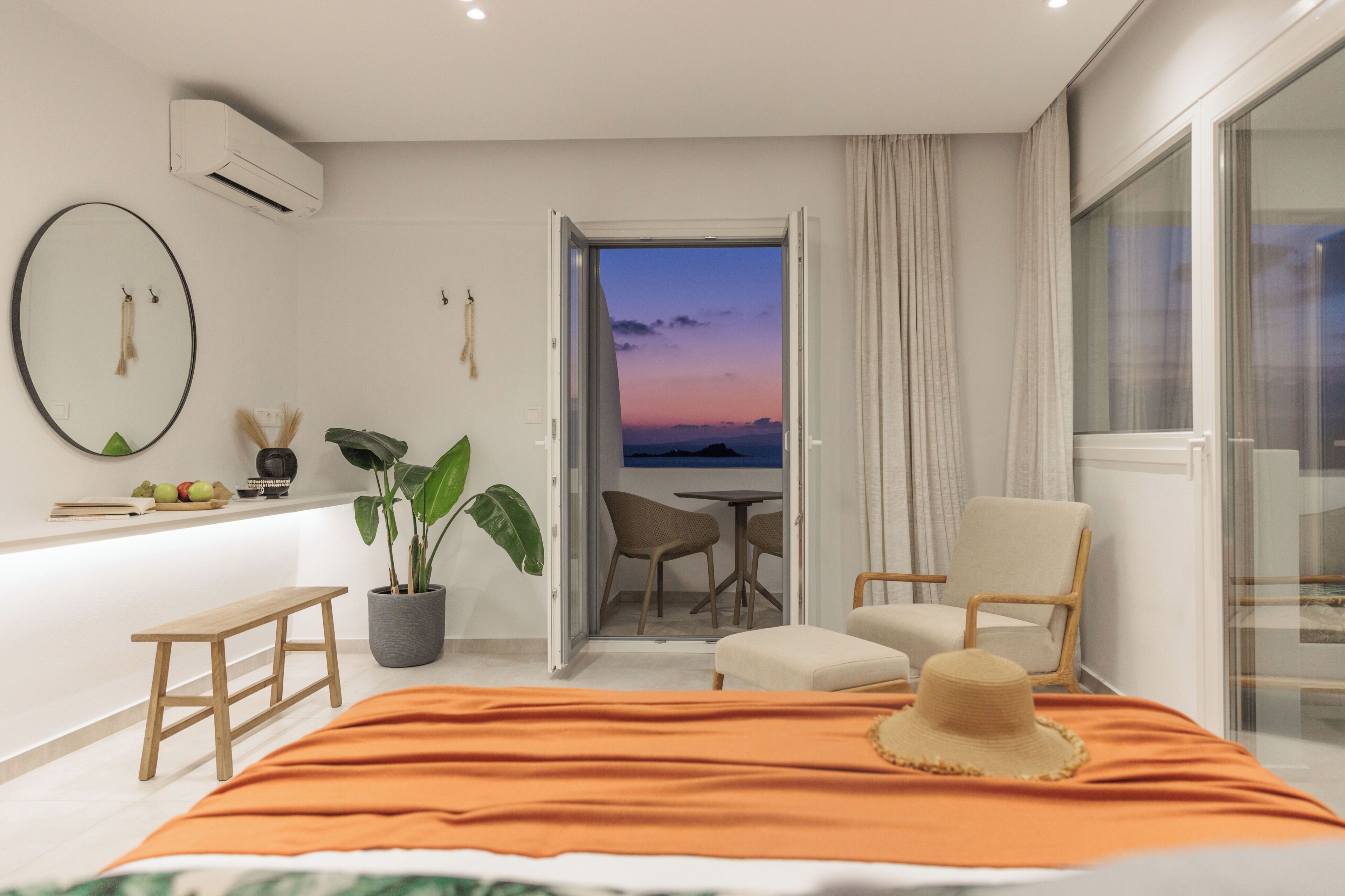 A modern hotel room with a bed, a small table, a chair, a potted plant, and a view of the sunset through an open door.