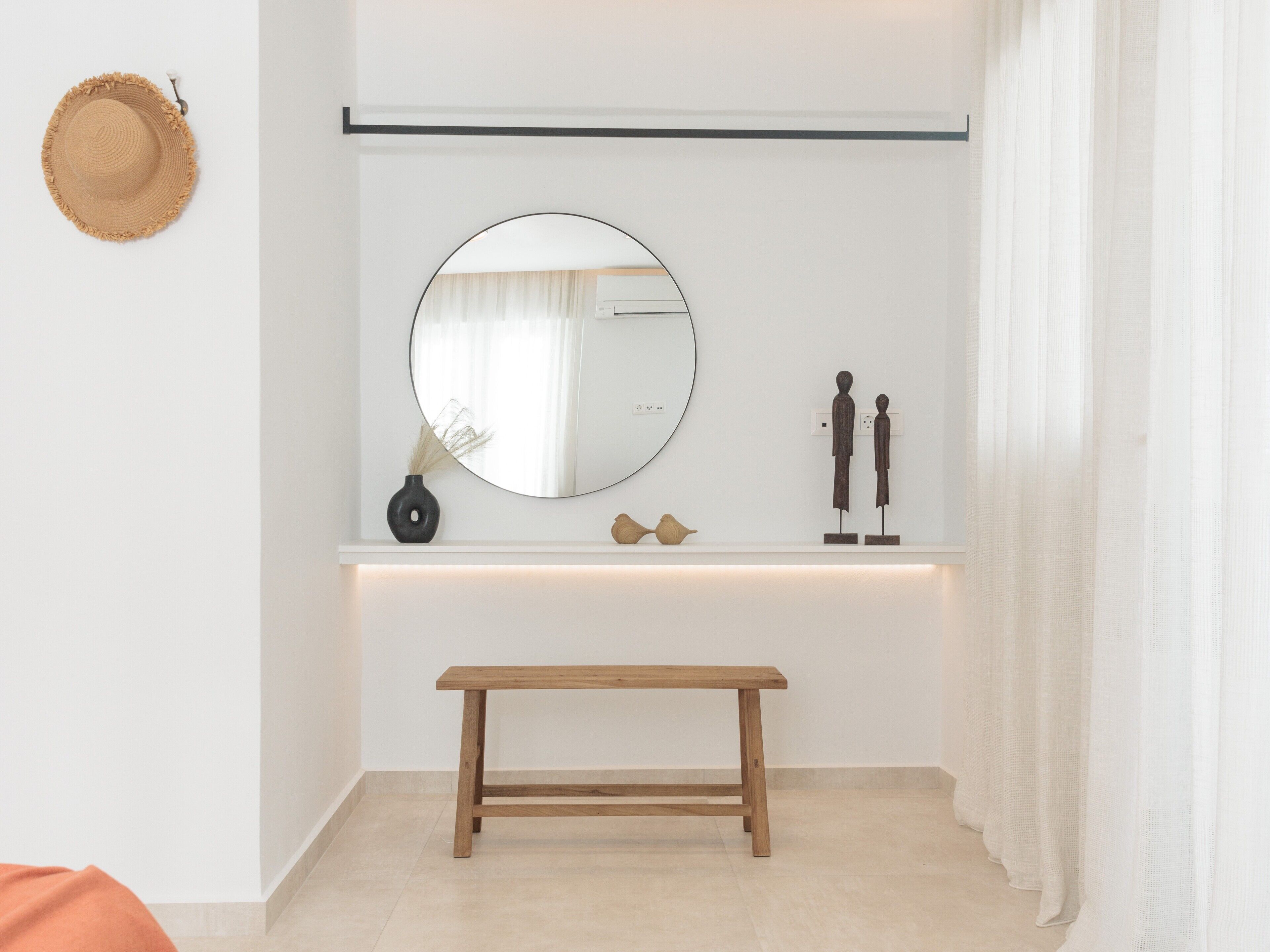 Una habitación minimalista con un banco de madera, un espejo redondo, un jarrón y dos figuras de madera en una estantería.