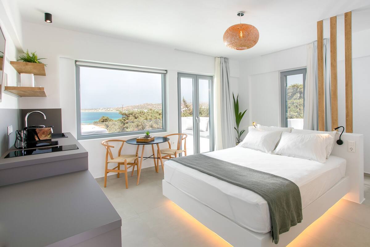 Un dormitorio moderno con una cama grande, una mesita pequeña y sillas, vista al mar y un balcón.