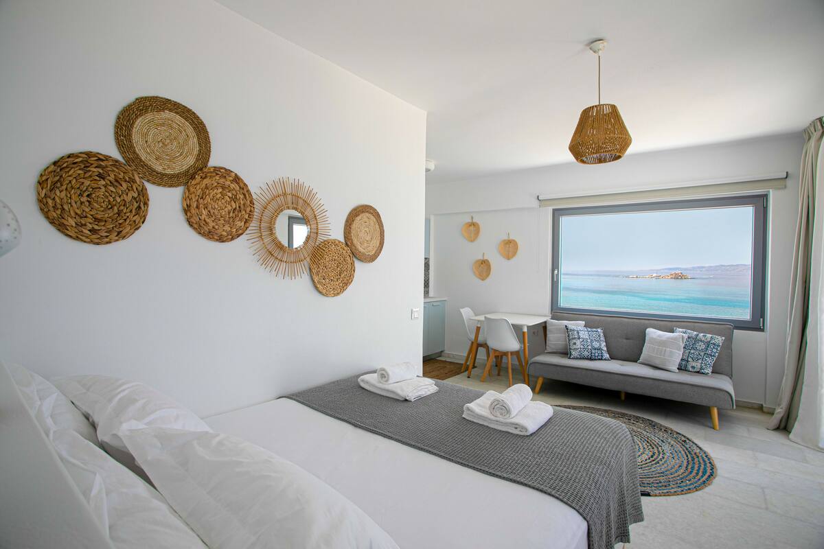 Habitación de hotel moderna con una cama grande, un sofá gris, zona de comedor y vistas al mar.