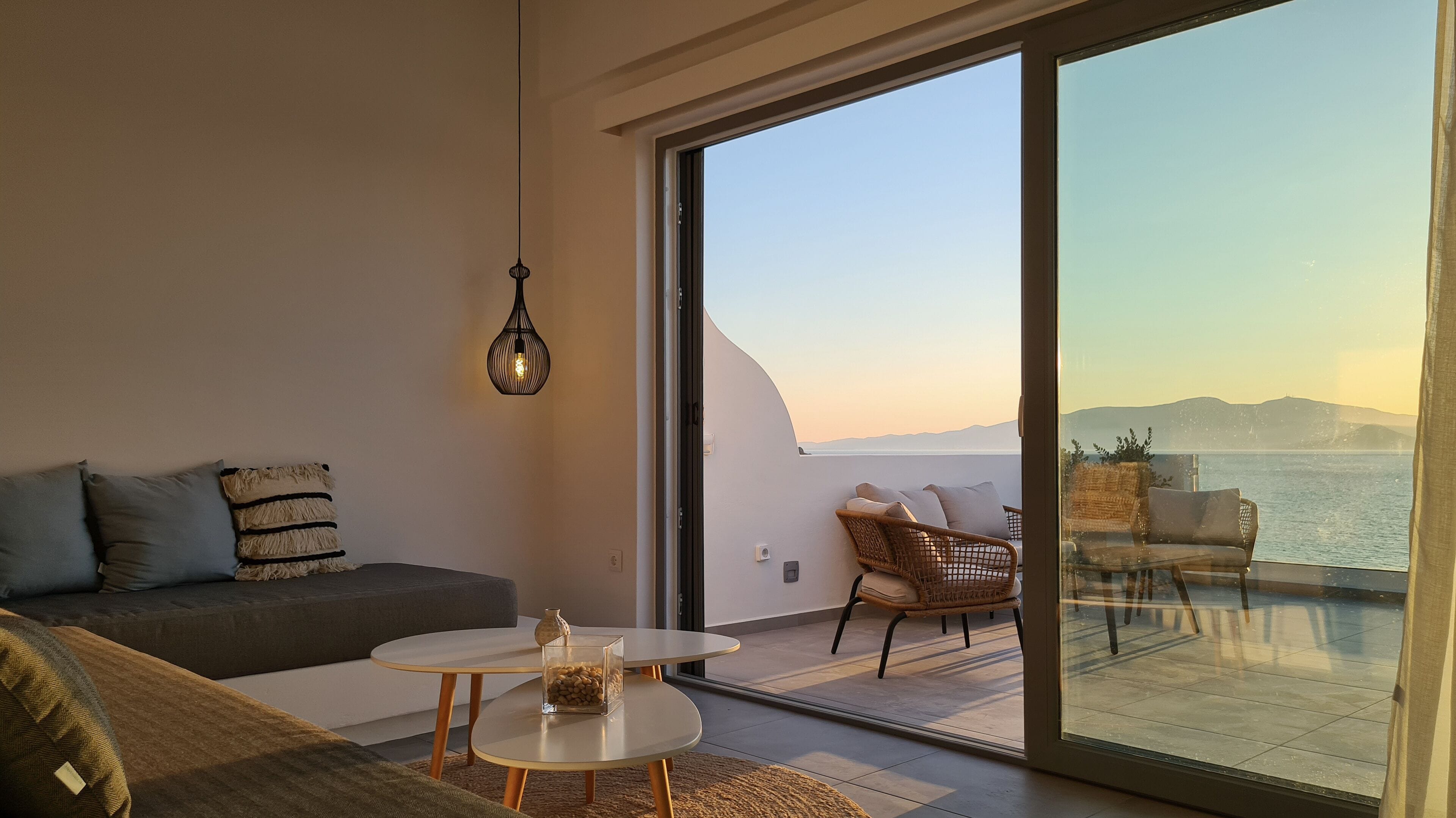 A modern living room with a sofa, a small table, and a wicker chair. A large sliding glass door leads to an outdoor balcony with a view of the sea and mountains.