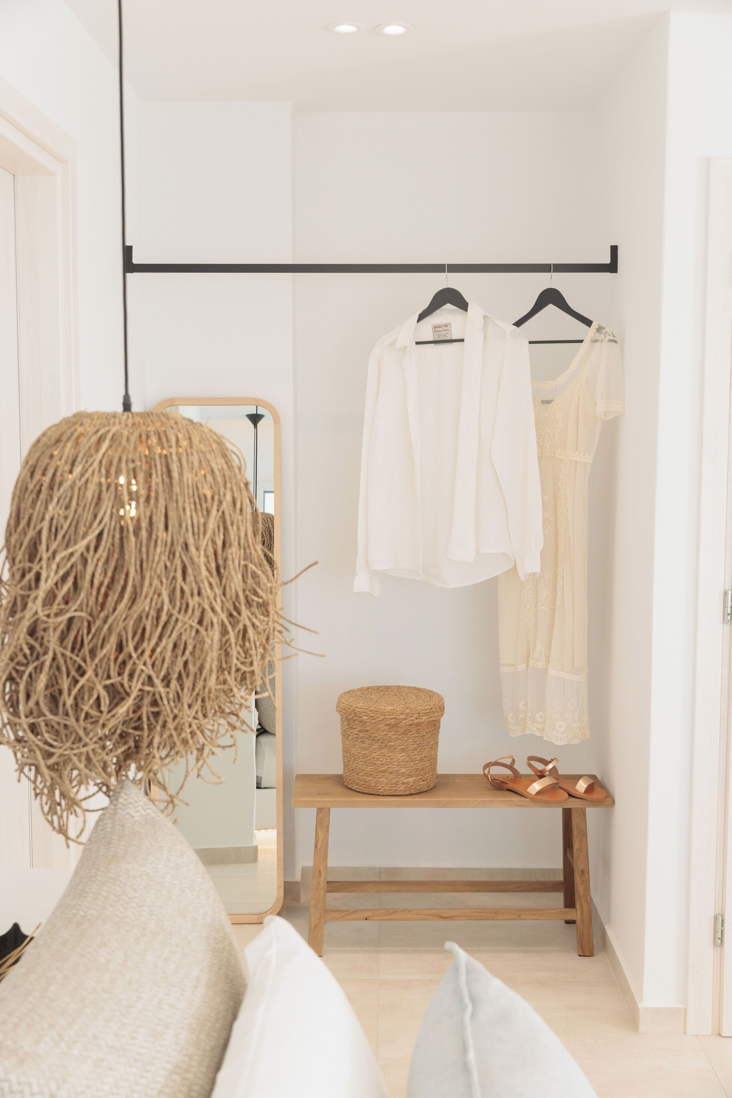 A room with a mirror, a hanging light fixture, a bench with a basket, and a clothing rack with a dress.