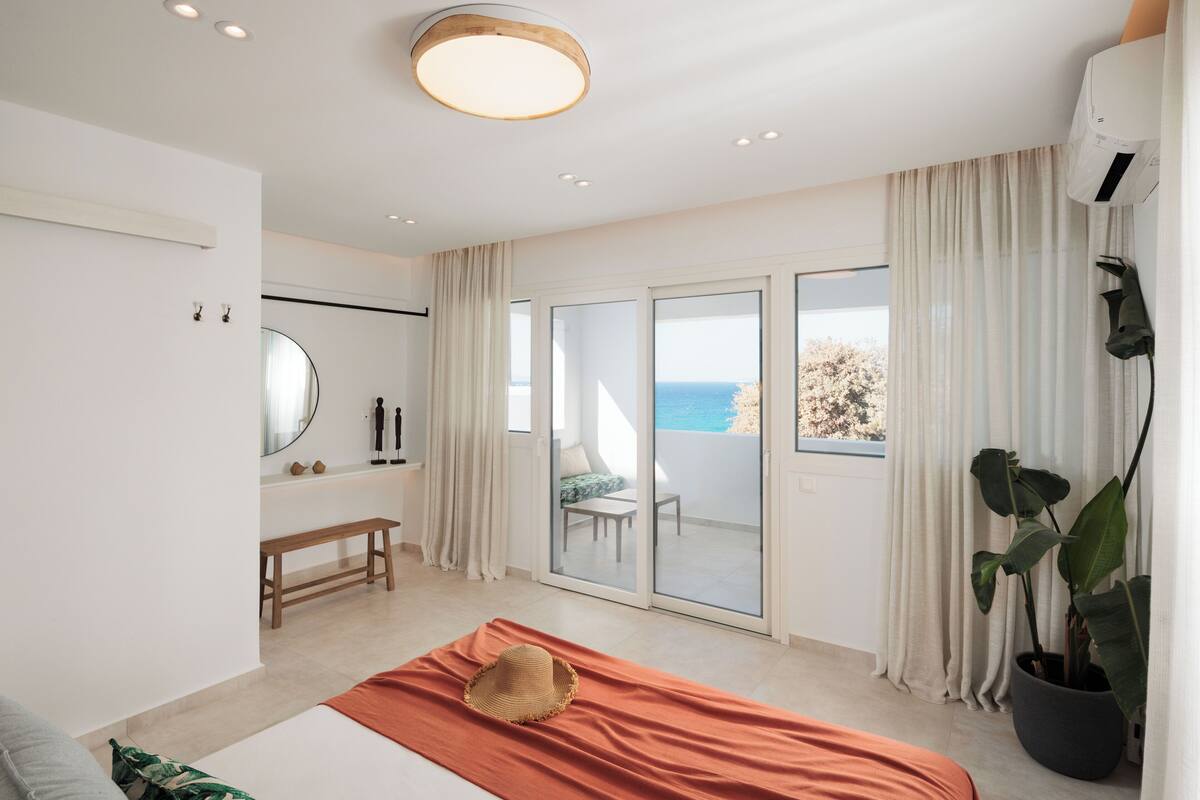 Un dormitorio moderno con cama, un banco, un ventanal con vista al mar y una planta en maceta.
