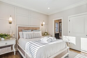 A bedroom with a large bed, white walls, a wooden headboard, two bedside tables with lamps, a nightstand with a plant, and a door with a window.