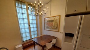 A dining area with a wooden table, white chairs, a chandelier, a large window with curtains, a refrigerator, and a framed map on the wall. - Italian designed apartment Marais (Paris)