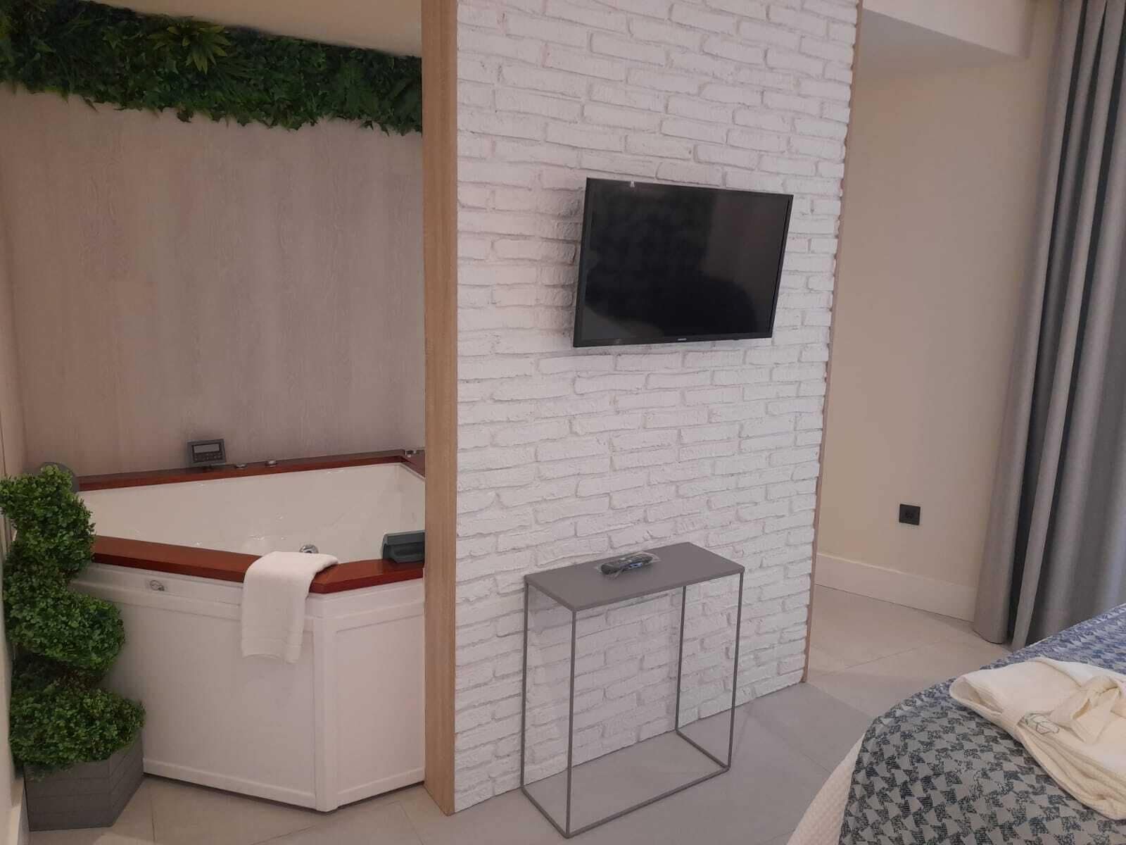 A modern bathroom with a white brick wall, a large bathtub, a small table, and a flat-screen TV mounted above the wall.
