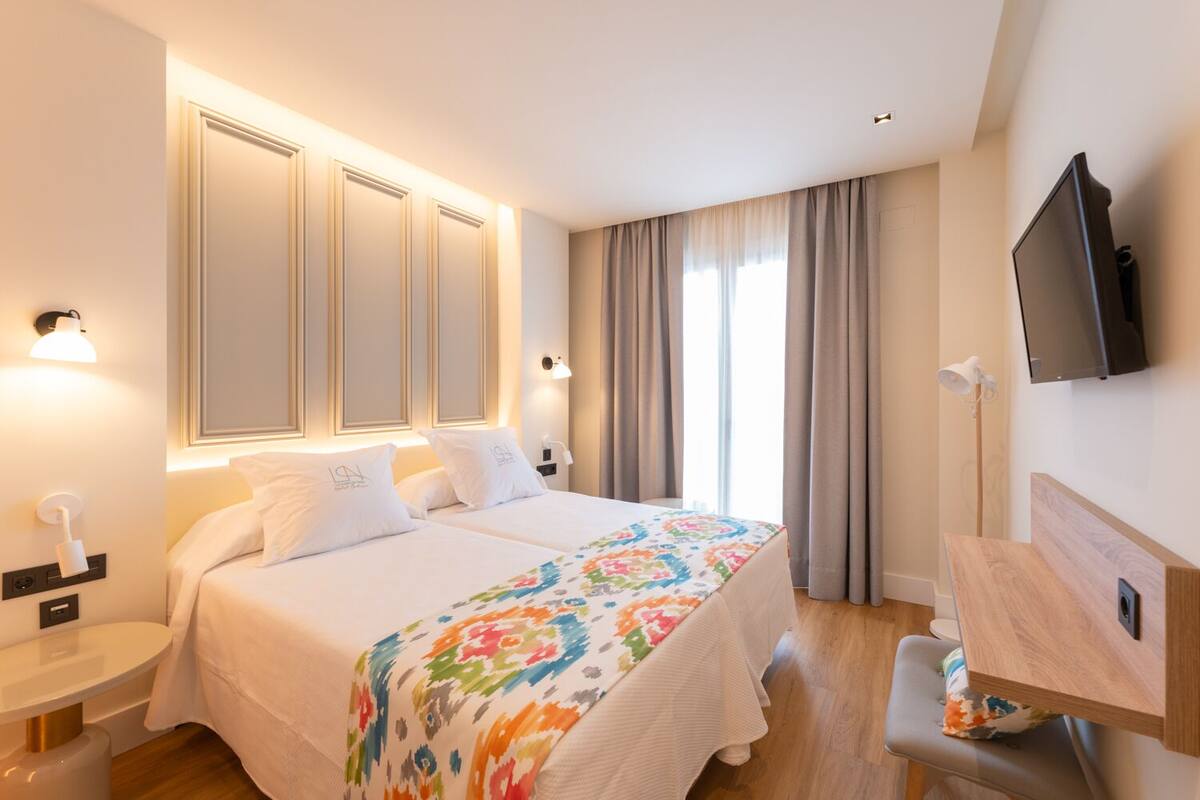 Habitació d'hotel amb un llit gran, dues taules de nit, un escriptori, una televisió i una finestra amb cortines.
