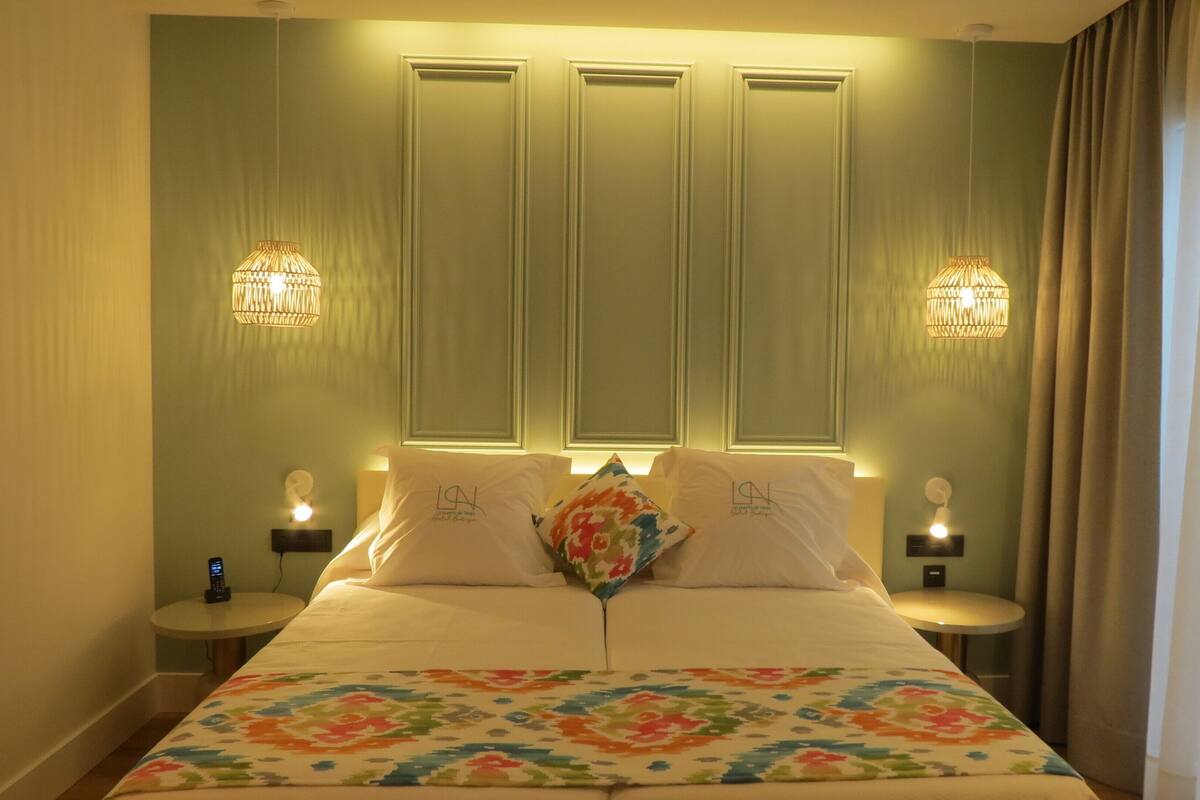 Habitació d'hotel amb un llit, dues taules de nit, una tauleta auxiliar, un coixí decoratiu i làmpades penjades a la paret.