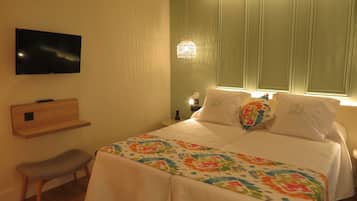 Ein Hotelzimmer mit Bett, Wandfernseher, Nachttisch mit Lampe und einer kleinen Holzablage.