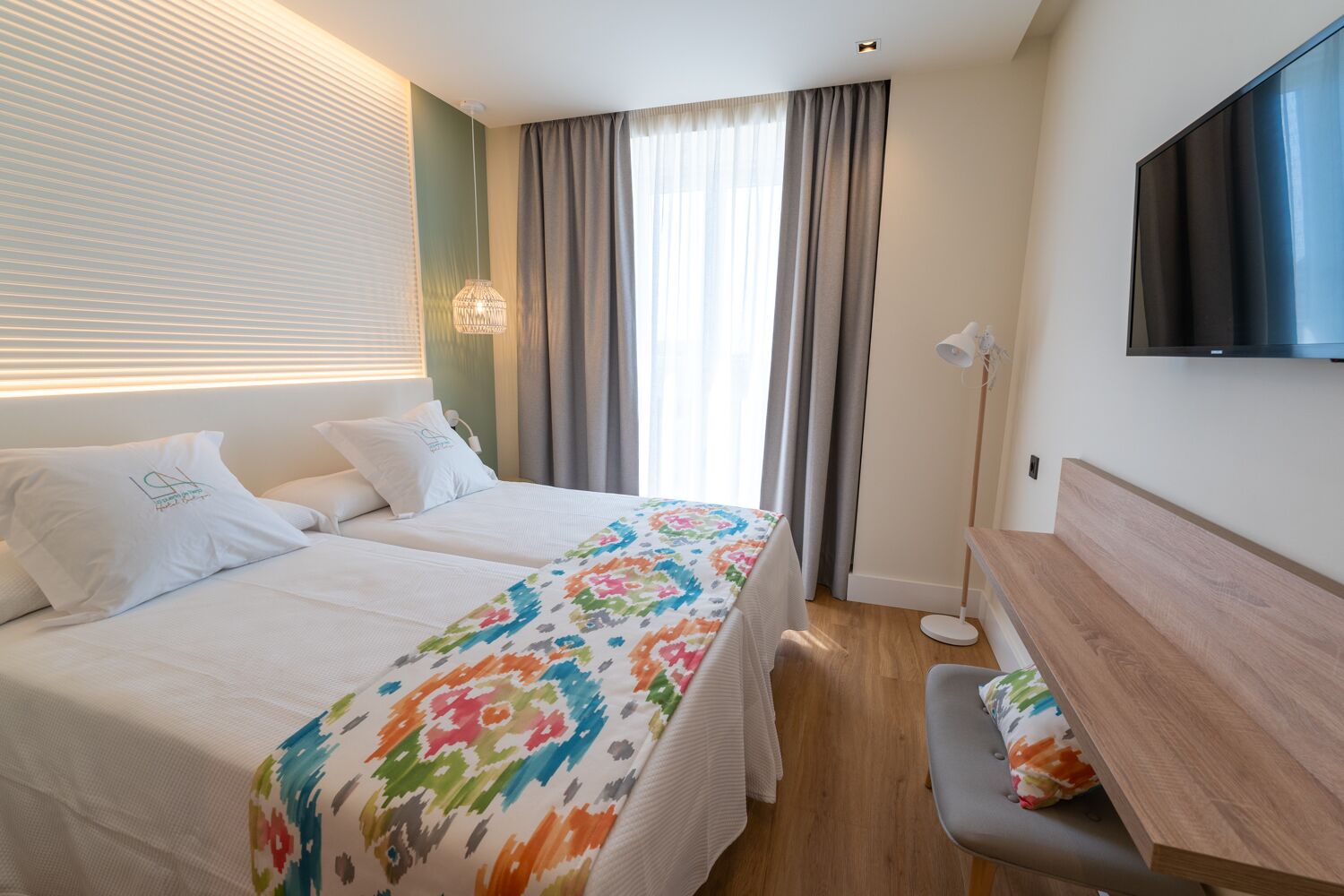 A hotel room with two beds, a desk, a TV, and a window with curtains.