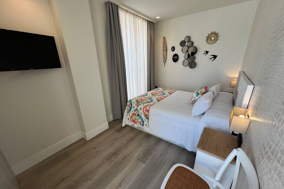 Habitació d'hotel amb un llit, una cadira, un televisor fixat a la paret i una finestra amb cortines.