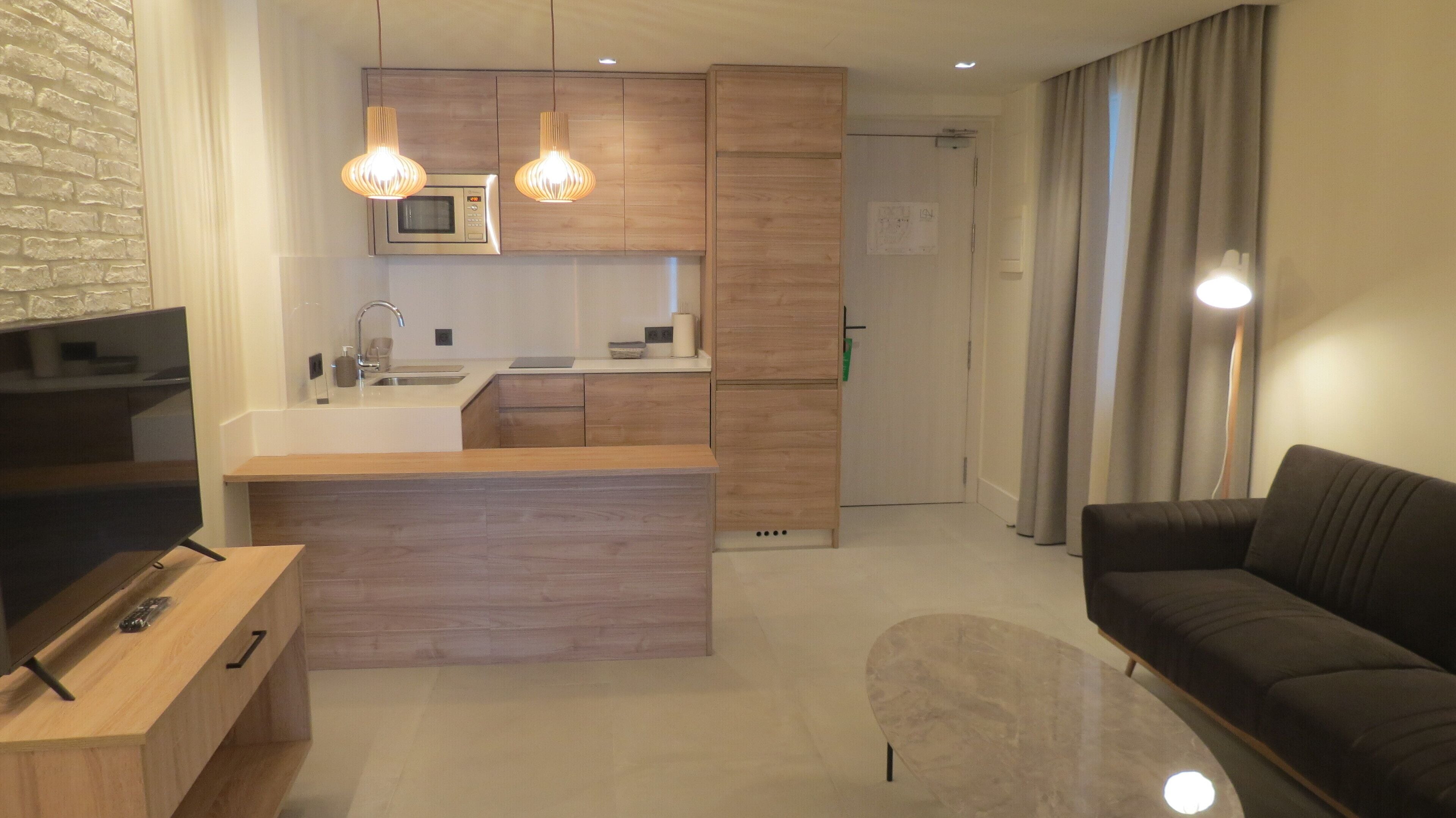 A modern living room with a dark sofa, a wooden coffee table, and a TV. A kitchen area with a microwave and wooden cabinets is visible in the background.