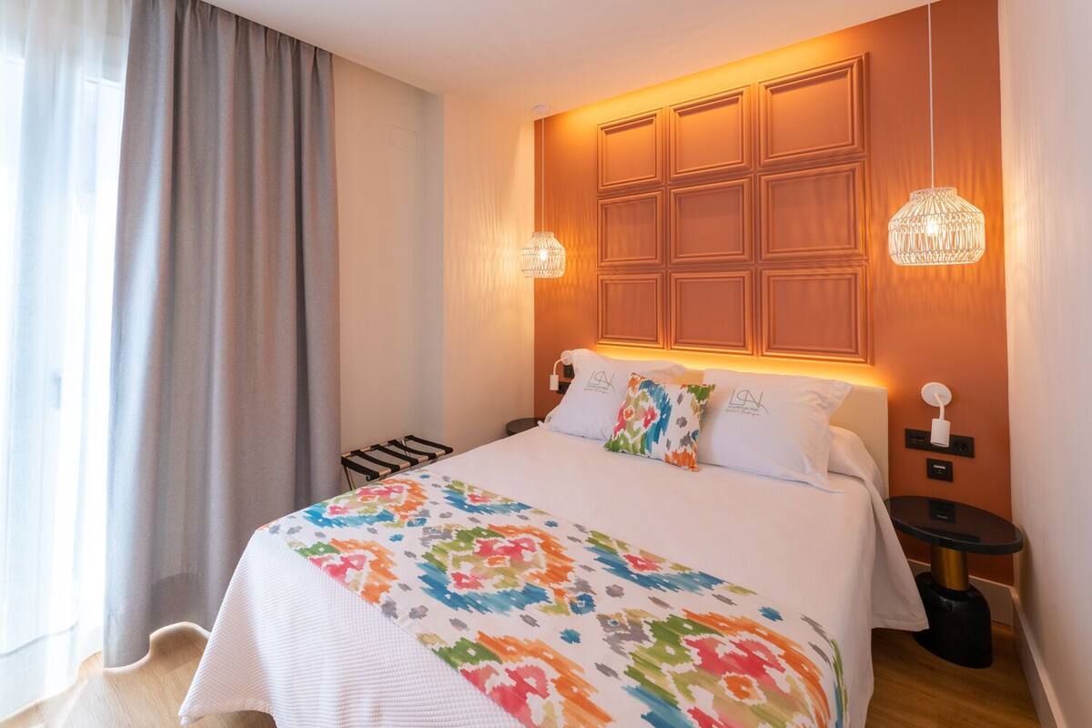 Habitació d'hotel amb un llit, una tauleta de nit, una làmpada i una finestra amb cortines.