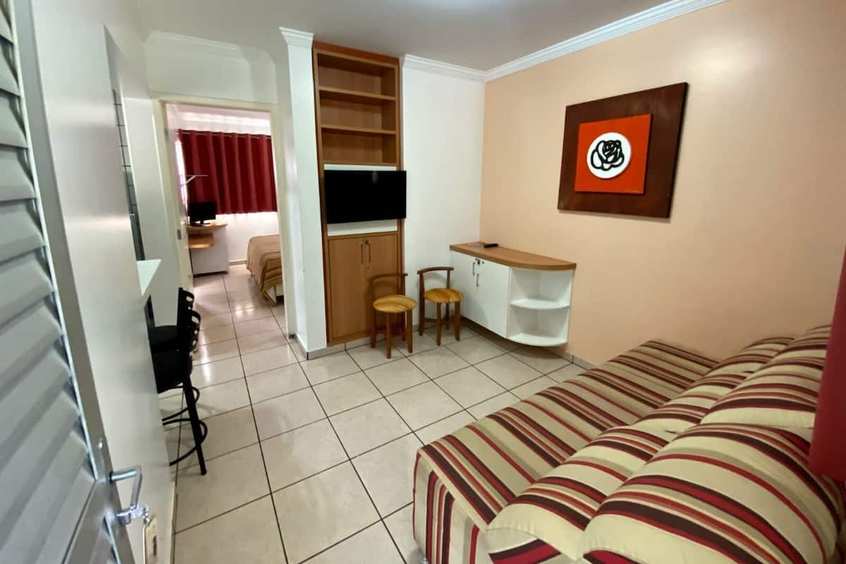 Un espace de vie compact comprenant un canapé, un téléviseur fixé au mur, un petit bureau avec une chaise et une bibliothèque.