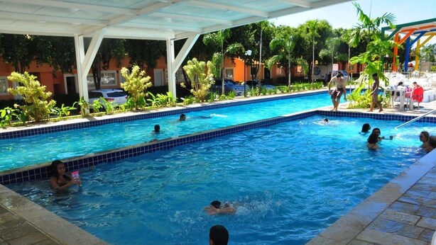 A swimming pool with people in the water, surrounded by greenery and a covered area. - Diroma Fiori - JC TEMPORADA (Caldas Novas)