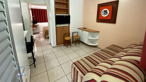 A compact living space with a sofa, a TV mounted on a wall, a small desk with a chair, and a bookshelf. - Diroma Fiori - JC TEMPORADA (Caldas Novas)