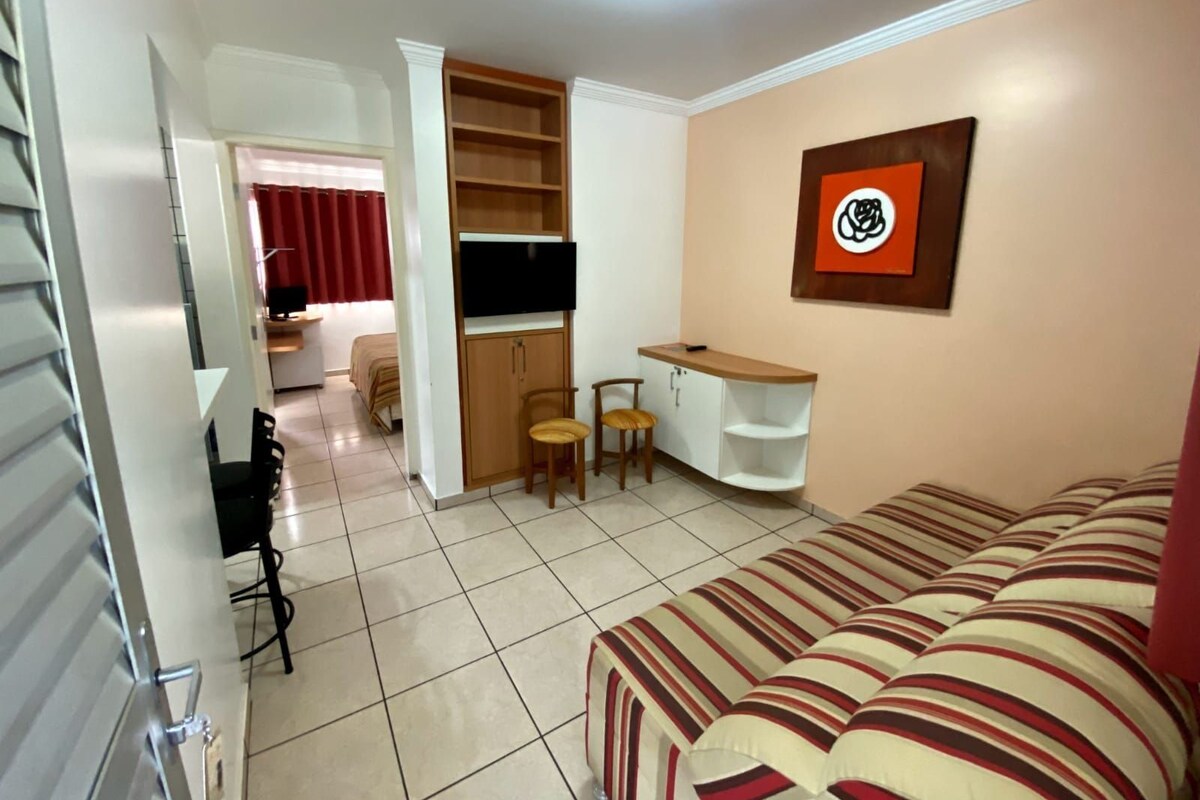 Ein kompakter Wohnraum mit Sofa, Wand-Fernseher, kleinem Schreibtisch mit Stuhl und Bücherregal.
