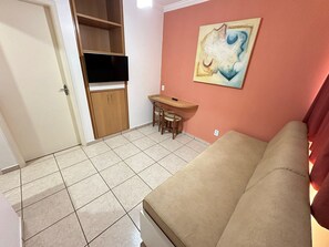 Ein Zimmer mit einem beigen Sofa, einem Holztisch mit zwei Stühlen, einem Fernseher auf einem Regal und einem Bild an der Wand.