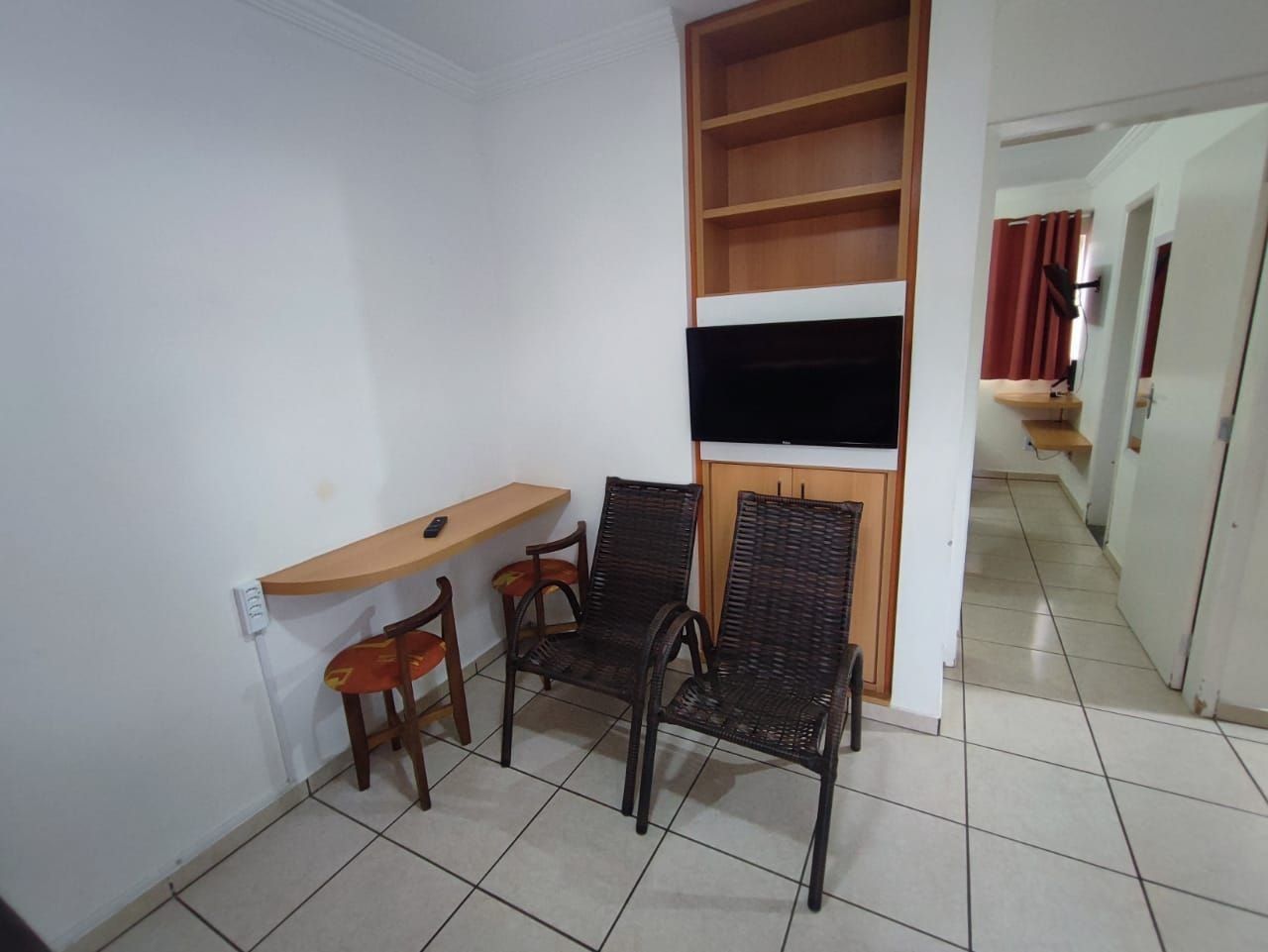 Una habitación pequeña con un escritorio de madera, dos sillas de mimbre, un televisor montado en un estante de madera y un piso de baldosas.