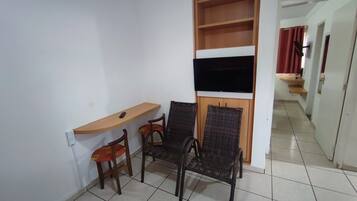Una habitación pequeña con un escritorio de madera, dos sillas de mimbre, un televisor montado en un estante de madera y un piso de baldosas.