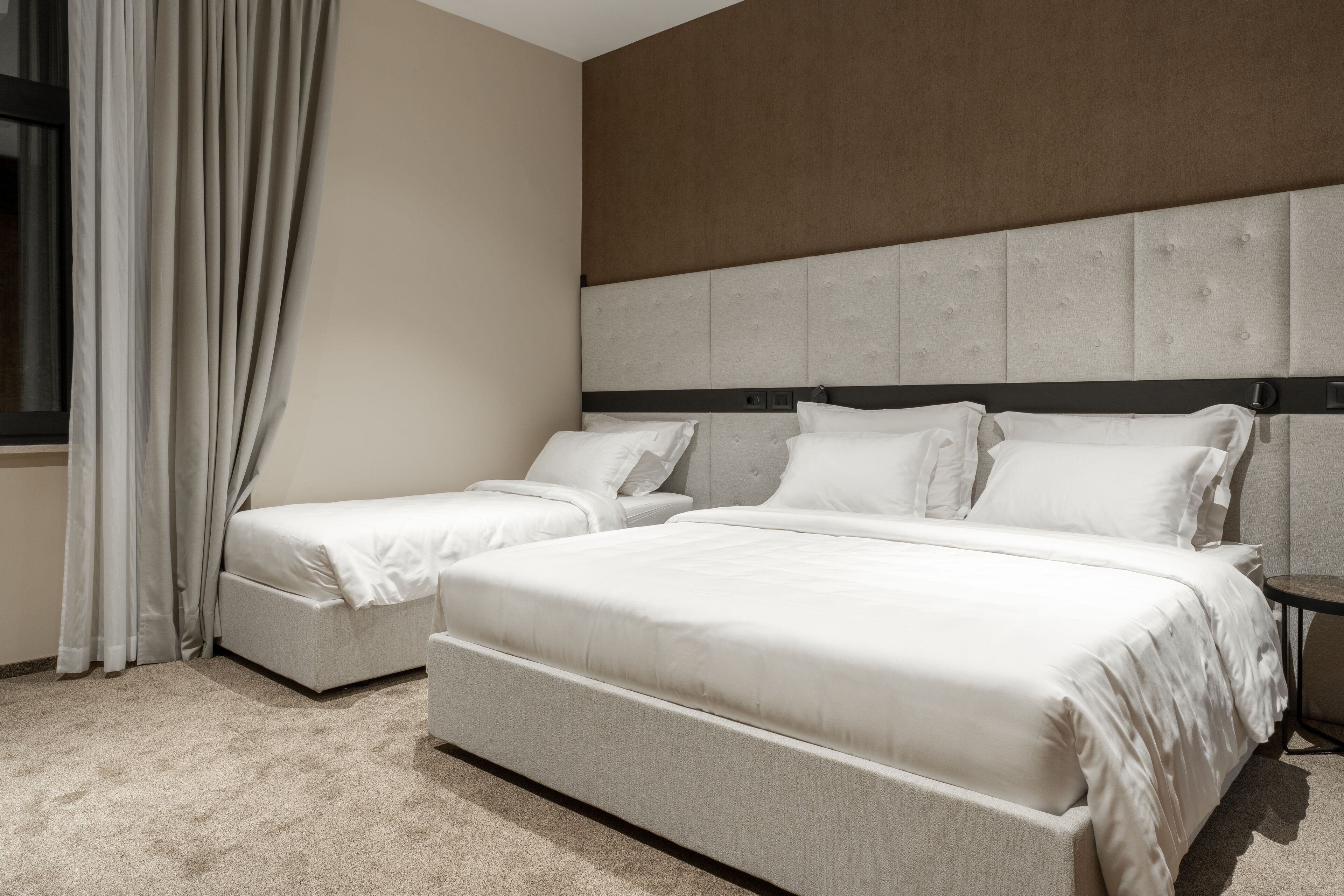 Una habitación de hotel con dos camas, un ventanal con cortinas y un cabecero con diseño acolchado.