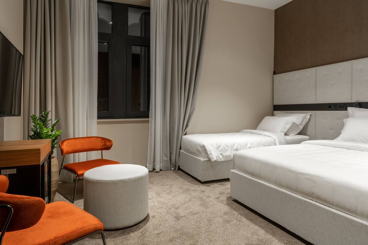 Quarto de hotel com duas camas, uma escrivaninha, uma cadeira e um banquinho redondo.