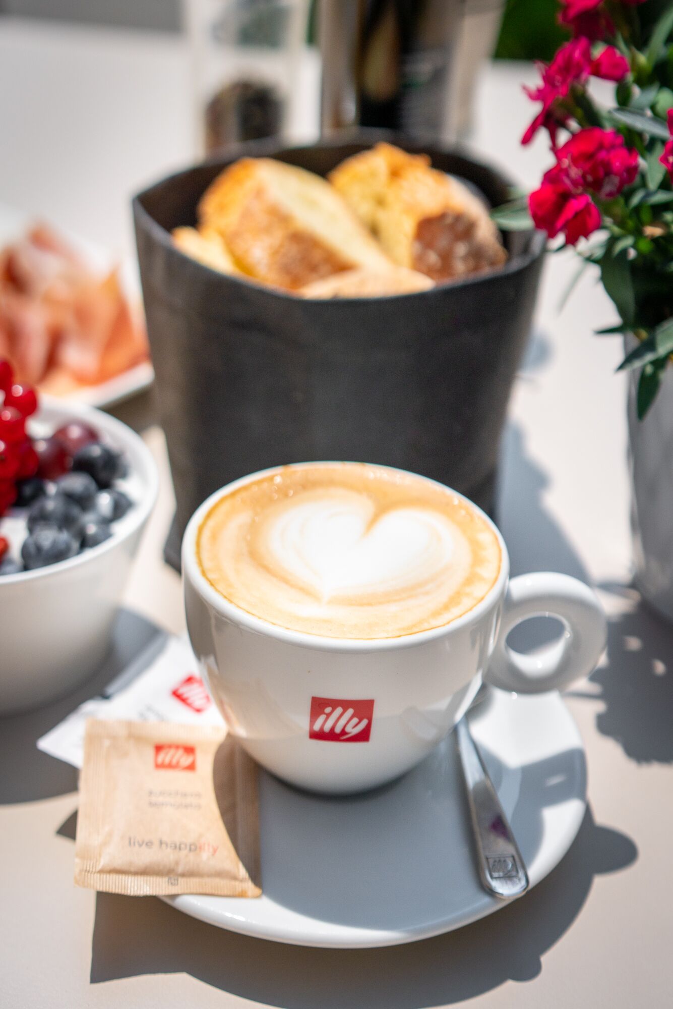 A cup of coffee with a heart-shaped latte art, a packet of Illy coffee, a bowl of berries, and a basket of pastries on a table.