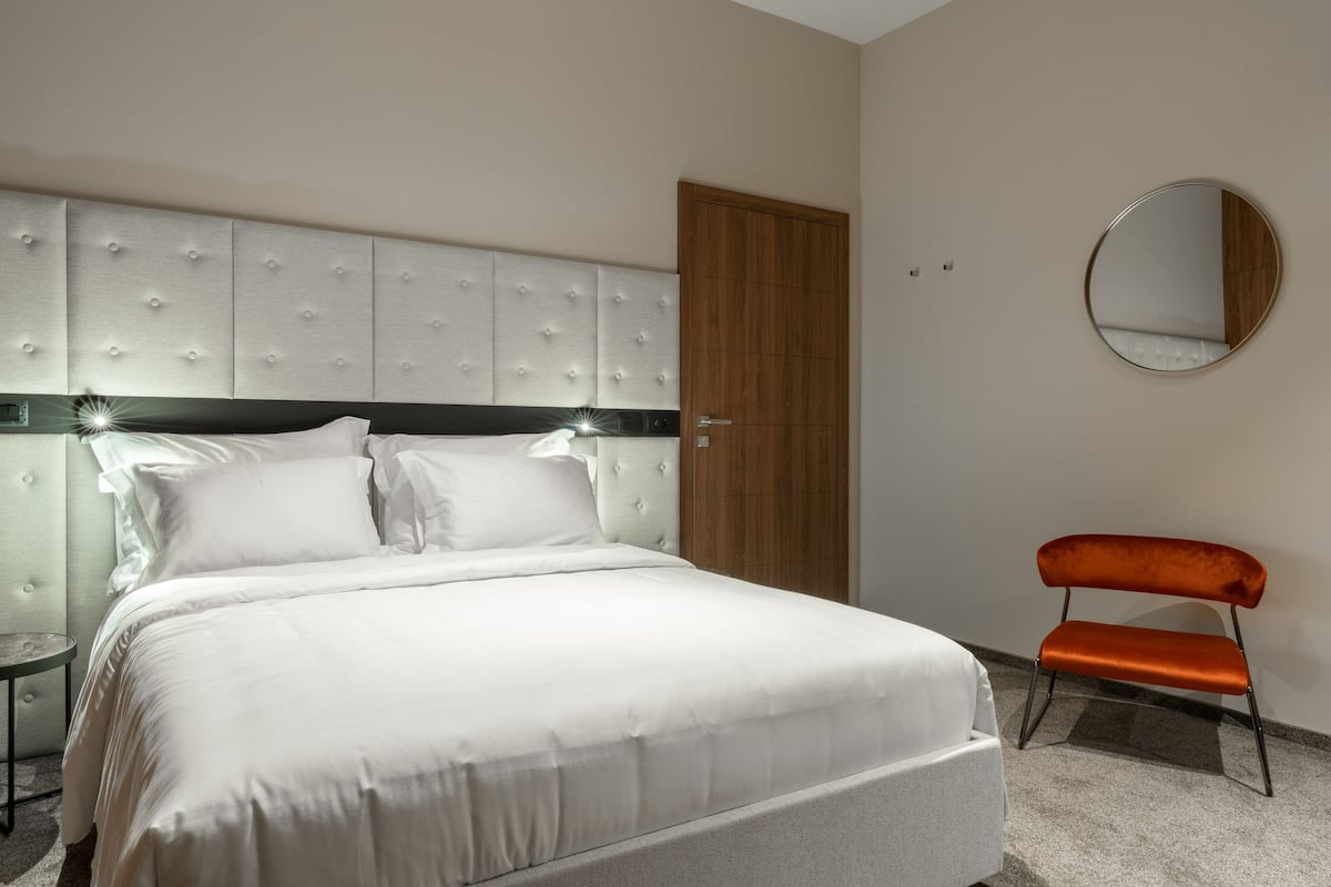 Uma cama bem arrumada com cabeceira estofada, uma porta de madeira e um espelho redondo na parede.