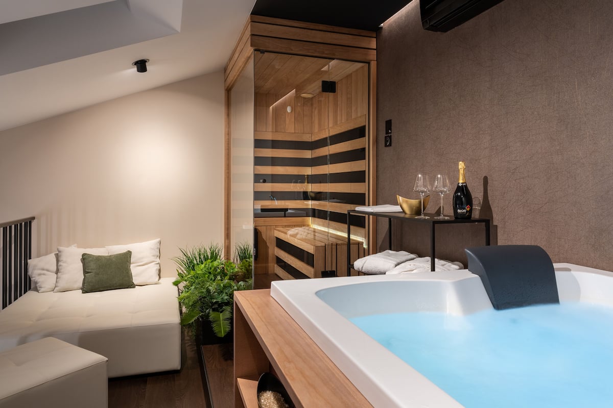 Um spa moderno com uma banheira grande, sauna de madeira e área de estar com sofá e uma mesinha com copos e garrafa.