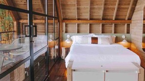 A wooden cabin bedroom with a large bed, a glass balcony, and a small table. - Cabaña Vacacional el Peñol con Jacuzzi (Guatapé)