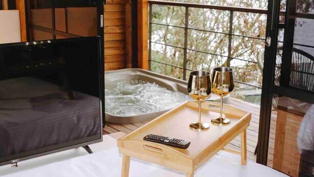 A cozy room with a bed, a hot tub, and a tray with two wine glasses and a remote control. - Cabaña Vacacional el Peñol con Jacuzzi (Guatapé)