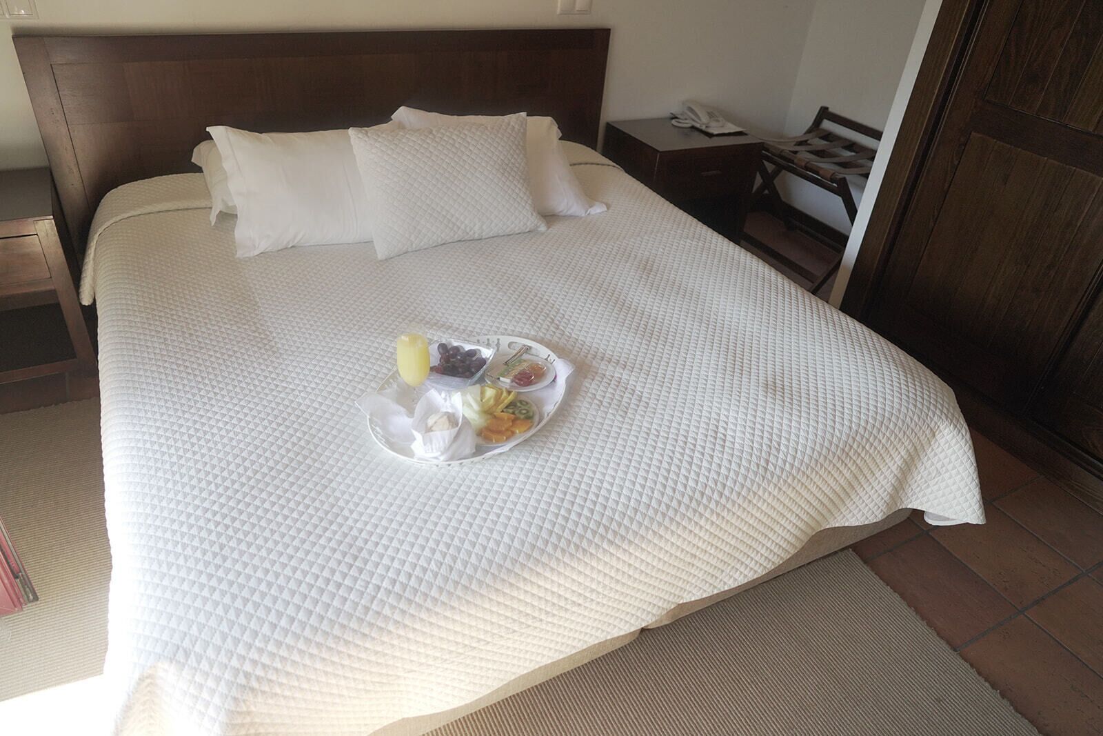 A neatly made bed with a white quilt, a tray of breakfast items including fruits and a drink, and a wooden nightstand with a phone.