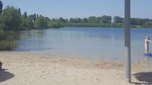 A beach with a thatched umbrella, sandy shore, calm water, and greenery in the background. - "Little uncle" (Wiefelstede)