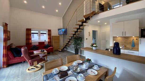 A modern living room with a red sofa, a dining table set with plates and cups, and a staircase leading to an upper level. - "Little uncle" (Wiefelstede)