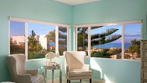 A room with a large window offering a view of a coastal landscape, a bed, two chairs, and a small table with a lamp. - Oceanview Luxury Home - 5 Bedroom - 5 Baths - 3 Decks - Walk to Beach (San Diego)