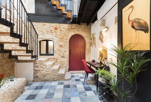 Una habitación con paredes de piedra, suelo a cuadros, una puerta roja, una escalera y un comedor con mesa y sillas.