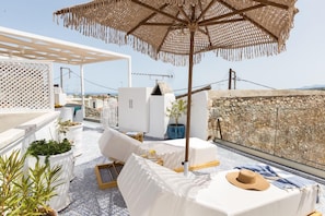 Terraza en la azotea con mobiliario blanco, una sombrilla grande de fibras naturales y plantas en macetas.