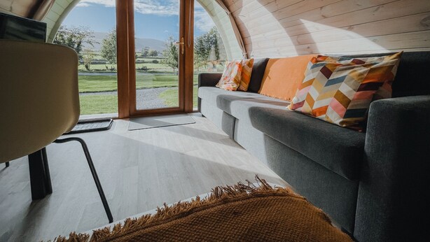 A cozy room with a curved wooden ceiling, a grey sofa with patterned cushions, a wooden floor, and a large window offering a view of a green landscape. - Comeragh Pods (Comeragh)