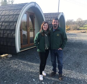 A couple standing in front of a small, curved-roofed structure with a glass door, set on a gravel surface.