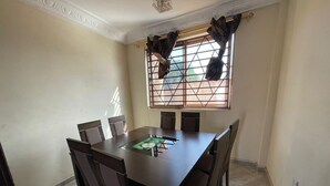 A dining room with a rectangular table and six chairs, a window with patterned curtains, and a decorative ceiling. - Cozy Kumasi Apartment With 2 Bedrooms (Kumasi)