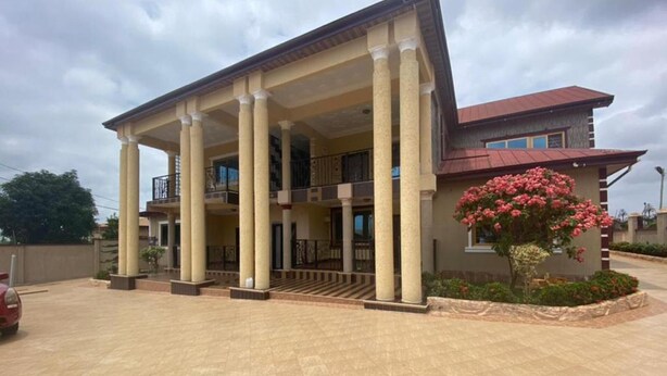 A building with a covered entrance, multiple columns, a balcony, and a red roof. - Cozy Kumasi Apartment With 2 Bedrooms (Kumasi)