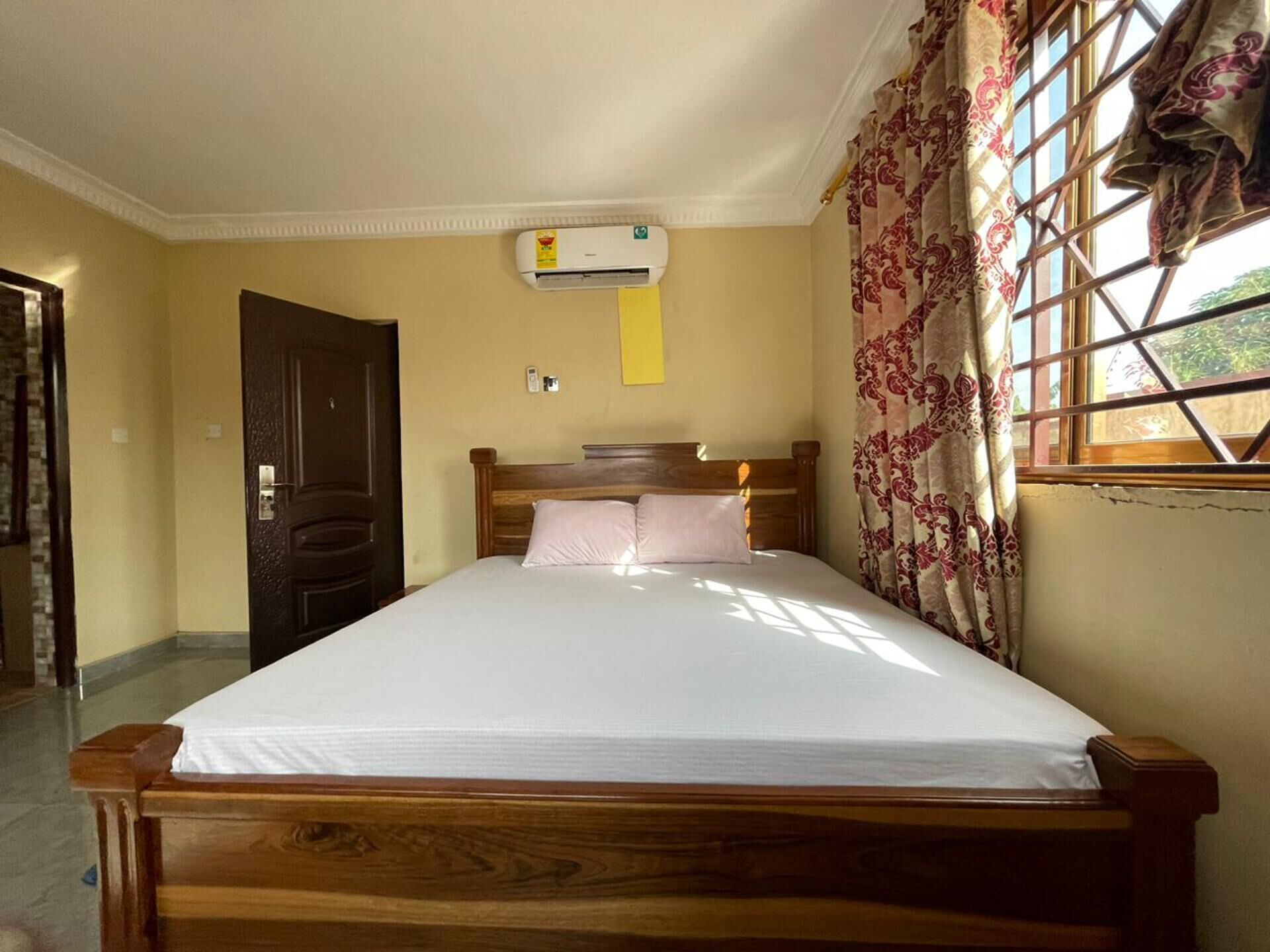 A room with a wooden bed, an air conditioning unit, a door, and a window with patterned curtains.