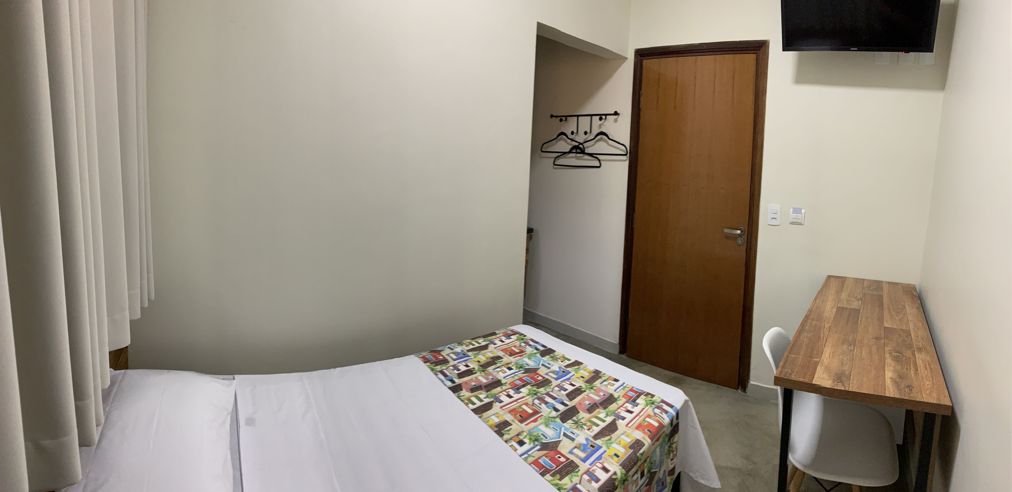A small, neatly arranged bedroom with a bed, a wooden table, a chair, a TV mounted on the wall, and a door.