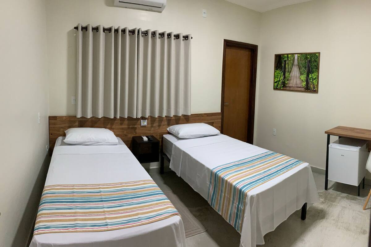 Una habitación de hotel con dos camas individuales, un cabecero de madera, un aire acondicionado blanco, una puerta de madera, un escritorio pequeño con una silla y un cuadro enmarcado en la pared.