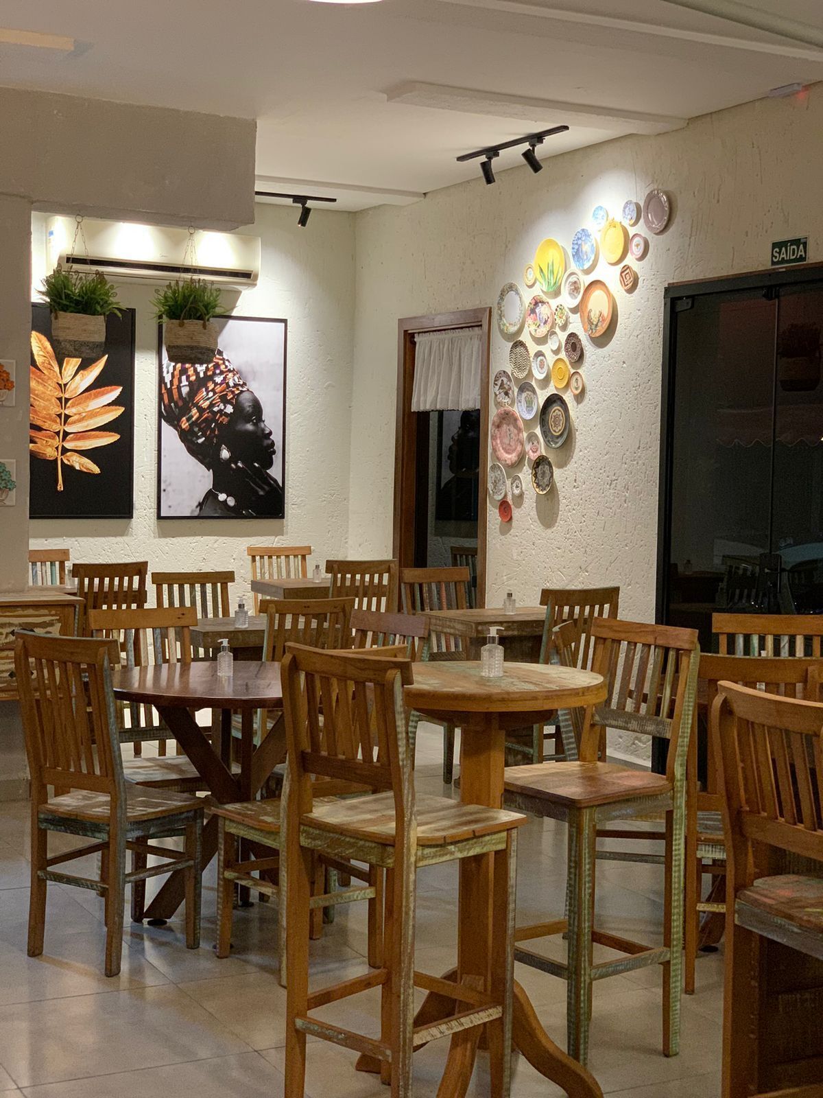 Ein Restaurantinnenraum mit Holztischen und -stühlen, an der Wand befestigten Tellern und zwei gerahmten Kunstwerken.