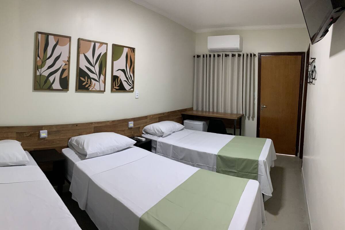 Habitación de hotel con tres camas individuales, cabecera de madera, mesita de noche, escritorio y aire acondicionado.