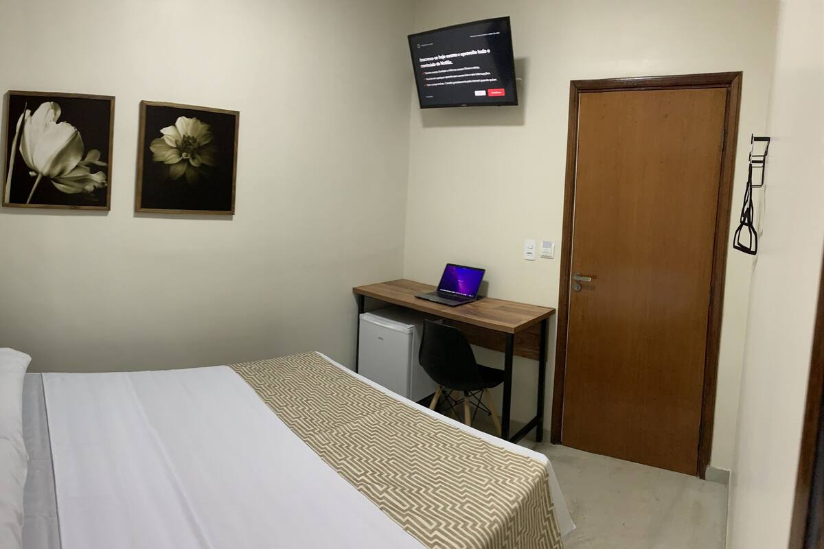 Un dormitorio con cama, escritorio con laptop, televisor y dos cuadros florales en la pared.