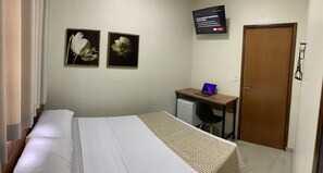 Ein Schlafzimmer mit einem Bett, einem Schreibtisch mit Laptop, einem Fernseher und zwei gerahmten floralen Bildern an der Wand.