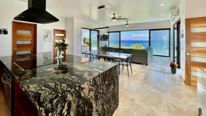 A modern kitchen with a marble countertop and a large island, a dining area with a view of the ocean, and a living area with a sofa and large windows.